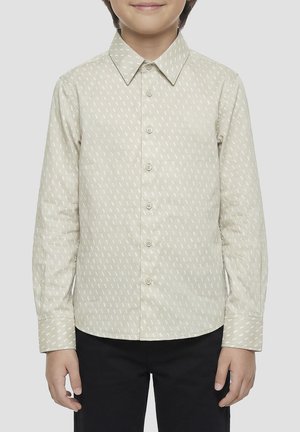 Kind draagt een beige overhemd met lange mouwen met een subtiel herhalend patroon, gecombineerd met een zwarte broek, staand tegen een effen achtergrond.