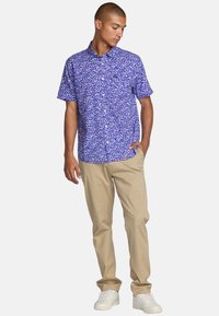 Short-sleeve shirt in blue with white floral pattern. Model pairs it with light beige trousers and white sneakers. Casual summer style.