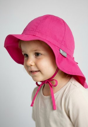 Toddler wearing a bright pink sun hat with chin ties, smiling and facing slightly left on a plain light background.
