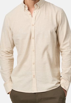 Homme portant une chemise beige à manches longues boutonnée avec un col boutonné et un pantalon marron, les mains dans les poches.