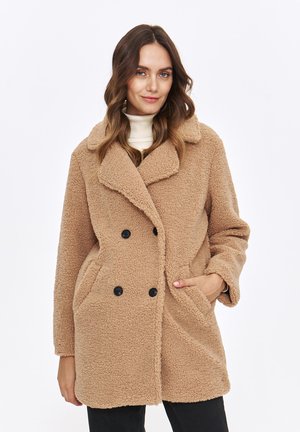 Beżowy, dwurzędowy płaszcz wykonany z polaru, z klapanym kołnierzem, dużymi kieszeniami i czarnymi guzikami. Miękka, fakturowana tkanina wykończeniowa.