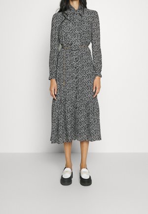 Vrouw draagt een zwart-witte bloemen midi-jurk met lange mouwen en een fijn strikriempje, gecombineerd met witte en zwarte platformschoenen.