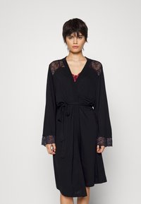 Robe noire à manches longues avec des accents en dentelle aux épaules et aux poignets. Taille cintrée, superposée sur un débardeur rouge. Texture de tissu lisse.