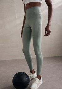 Leggings a vita alta, aderenti, in tessuto verde chiaro con superficie liscia. Accentuati da cuciture piatte, abbinati a scarpe da ginnastica bianche e a una palla nera.