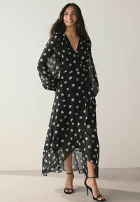 Rochie maxi neagră dintr-un material ușor și transparent, cu modele circulare albe. Are un decolteu adânc în V, mâneci lungi tip balon și un stil înfășurat.