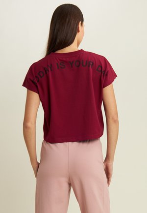T-shirt borgogna cropped a maniche corte, con la scritta nera "OGGI È IL TUO GIORNO" stampata sul retro. Abbinata a pantaloni rosa chiaro.