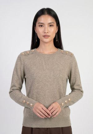 Frau mit langen schwarzen Haaren, die einen beigen Pullover mit dekorativen goldenen Knöpfen an Schultern und Ärmeln trägt, steht vor einem schlichten Hintergrund.