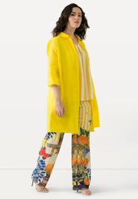 Camicia lunga gialla in lino sovrapposta a una maglietta a righe e pantaloni a gamba larga con motivi floreali e di frutta colorati. Scarpe con punta scoperta visibili.