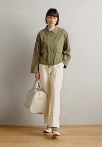 Olive green cropped jacket with two front pockets, paired with cream wide-leg pants and grey sneakers. Model carries a cream bag.
