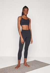 Mulher sorridente em pé descalça numa esteira de yoga salpicada, vestindo um top desportivo preto e leggings cropped pretas de cintura alta.