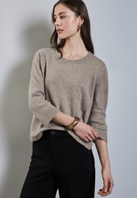 Beige gebreide trui met ronde halslijn en geribbelde manchetten, gecombineerd met zwarte broeken. Opvallende armband met gouden accenten om de pols.