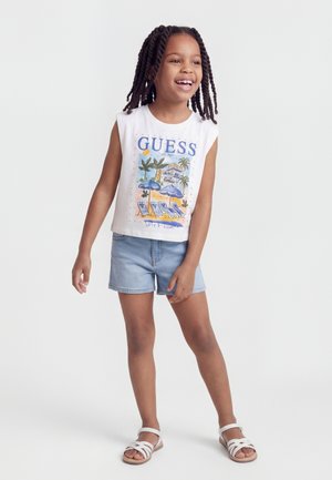 Lächendes junges Mädchen mit geflochtenem Haar, das ein weißes ärmelloses Oberteil mit Strandmotiv, Jeansshorts und weiße Sandalen trägt, vor weißem Hintergrund stehend.