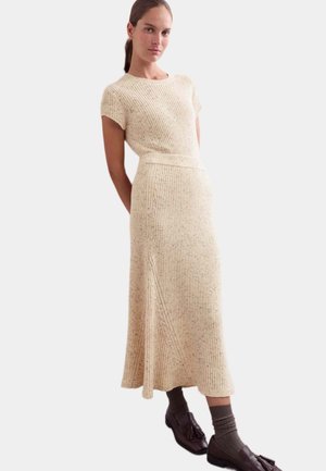 Donna che indossa un vestito in maglia a coste color crema con maniche corte e gonna midi, abbinato a calzini marroni e mocassini con nappine.