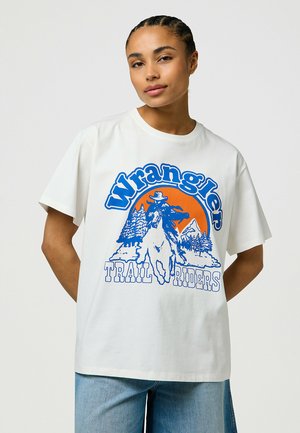 T-shirt en coton blanc avec un design graphique bleu portant le texte "Wrangler TRAIL RIDERS," représentant un cowboy sur un cheval devant des montagnes.