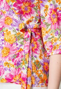 Blouse à encolure fleurie en tissu léger, présentant des fleurs roses, violettes et orange sur un fond blanc, avec un détail de taille nouée.