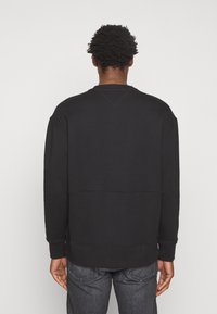 Uomo con capelli corti e ricci, vestito con una felpa nera a maniche lunghe semplice e jeans grigio scuro, rivolto di spalle su uno sfondo bianco.