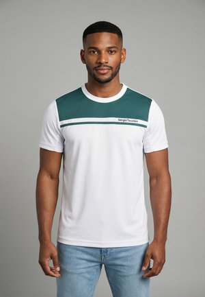 Uomo in piedi, rivolto in avanti, che indossa una camicia bianca a maniche corte con un pannello verde e jeans azzurri chiari su uno sfondo grigio.