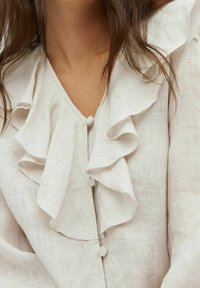 Blusa di lino con scollo arricciato, tessuto leggero, colore crema e chiusure frontali con bottoni. Inquadratura ravvicinata sui dettagli.