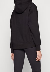 Personne portant un sweat à capuche noir uni et des leggings noirs, vue de dos avec une main détendue le long du corps.