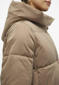 Chaqueta acolchada beige con un alto cuello y capucha. Fabricada con un material suave y ligero, con costuras visibles y un patrón acolchado.