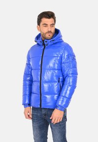 Giacca a vento blu con finitura lucida, chiusura frontale con zip, cappuccio, design imbottito e tasche laterali. Abbinata a jeans in denim scuro.