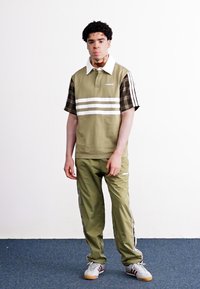 Olivengrünes Poloshirt mit karierten Ärmeln und weißen Streifen, kombiniert mit passenden olivgrünen Hosen. Hellgraue Sneaker mit Streifen.