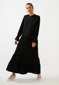 Nainen seisoo mustassa, pitkähihhaisessa, kerroksellisessa maksihaalarissa, jossa on edessä koristepitsi. Hän on yhdistänyt sen vaaleisiin lenkkareihin ja helmikorvaketjuihin.