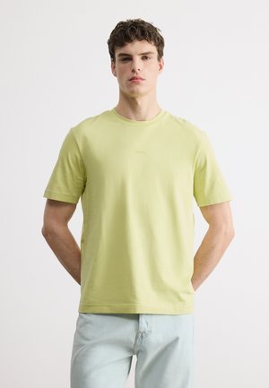 Młody mężczyzna stoi przodem, ubrany w jasnożółtą koszulkę z krótkim rękawem i jasnoniebieskie dżinsy na tle jednolitej białej ściany.