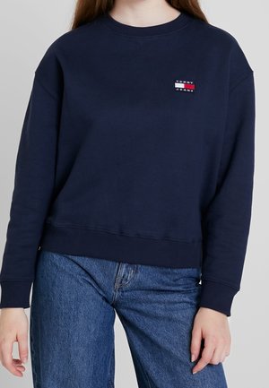 Persoon draagt een marineblauwe trui met een Tommy Jeans-logo op de borst en blauwe spijkerbroek, tegen een effen achtergrond.