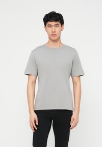 Jeune homme aux cheveux courts noirs portant un T-shirt gris clair uni à manches courtes et un pantalon noir, debout devant un fond blanc.
