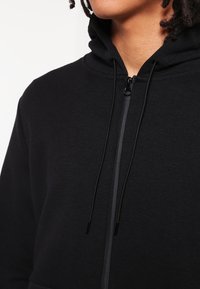 Felpa nera con zip, dotata di zip frontale, cappuccio regolabile con cordino e texture morbida. Il tessuto appare liscio e leggero.