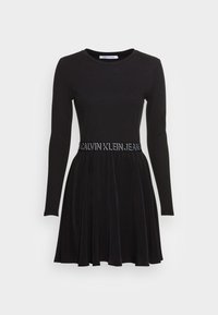 Black long-sleeve dress with a fitted top and flared skirt. Features a contrasting waistband with "CALVIN KLEIN JEANS" in white lettering.
