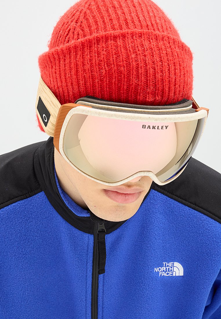 Skibrille mit einer geschwungenen, getönten Linse und einem beigen Rahmen, kombiniert mit einer roten Rippmütze und einer blauen Fleecejacke mit schwarzem Kragen.