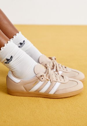 Stopy w beżowych butach Adidas Samba z białymi paskami oraz białych skarpetkach z logo Adidas na żółtej wykładzinie.
