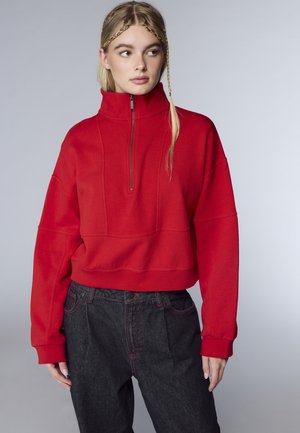 Giovane donna con lunghi capelli biondi e trecce indossa un maglione rosso a mezza zip e jeans neri a vita alta, guardando verso sinistra.