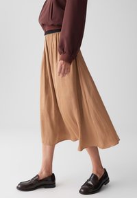 Una gonna fluida, di lunghezza al ginocchio, di colore beige chiaro, realizzata in un tessuto leggero, abbinata a una felpa marrone scuro, dal taglio comodo, e mocassini marroni.