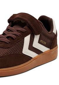 Brązowy zamszowy sneaker z fakturowaną cholewką, białymi akcentami w kształcie szewrona oraz paskiem na rzep. Posiada gumową podeszwę i przeszycia jako detale.