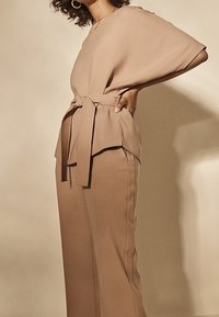 Kvinna som bär beige vida byxor och en matchande bälte topp med dolmansleeves, står med ena handen på höften mot en enfärgad bakgrund.