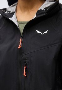 Μαύρο αδιάβροχο μπουφάν με γκρι φόδρα κουκούλας, φερμουάρ σε νέον πορτοκαλί χρώμα και λευκό γεωμετρικό λογότυπο στο στήθος, φορεμένο πάνω από πράσινο τοπ.