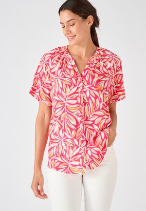 Mujer con una blusa de manga corta de color rosa brillante con patrones de hojas blancas y naranjas, combinada con pantalones blancos, sonriendo y mirando hacia abajo.