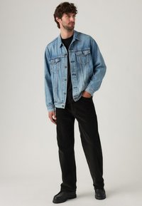 Man staat met een lichtblauwe spijkerjack, zwart shirt, zwarte broek en zwarte schoenen, met een hand in zijn zak en kijkt naar rechts.
