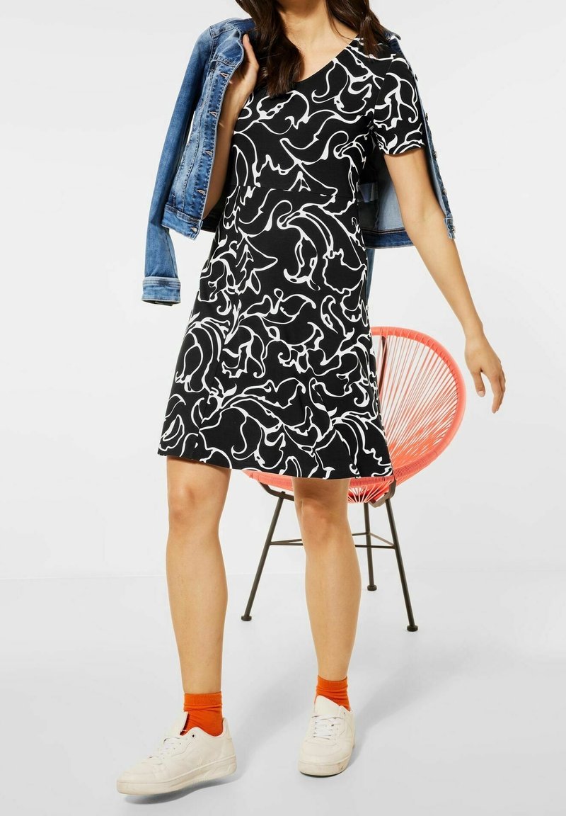 Robe noire avec des motifs blancs en spirale, manches courtes et jupe évasée, associée à une veste en denim, des chaussettes orange et des baskets blanches.