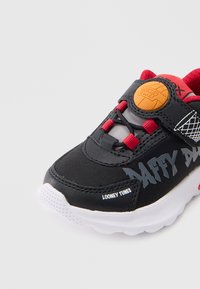 Ténis preto com detalhes em vermelho, estampado com o Pato Donald em cinzento, sola branca, parte superior texturizada e um logótipo circular laranja na tira.