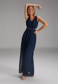 Navyblaues Maxikleid mit ärmellosem Design, ausgeschnittenem Taillenbereich, fließendem Stoff und einem taillierten Gürtel. Abgerundet mit glitzernden Sandalen.