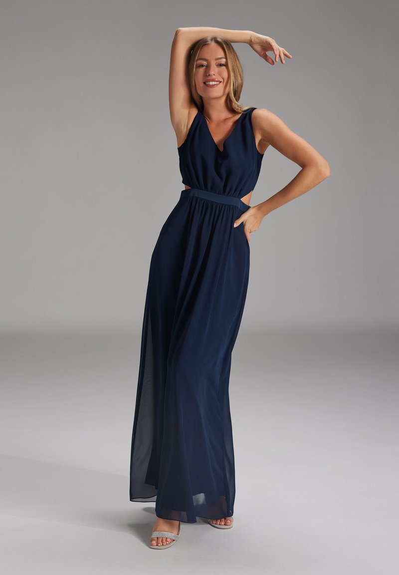 Navyblaues Maxikleid mit ärmellosem Design, ausgeschnittenem Taillenbereich, fließendem Stoff und einem taillierten Gürtel. Abgerundet mit glitzernden Sandalen.