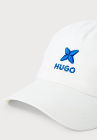 White baseball cap made of fabric featuring blue embroidered logo and emblem on the front. Soft texture with stitched detailing on the peak.