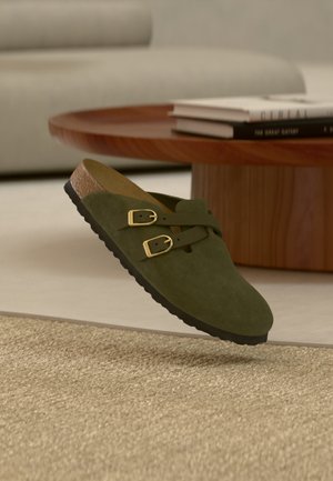 Grøn ruskind slip-on sandal med dobbelte gyldne spænder svæver over et beige vævet tæppe med et rundt træbord og bøger i baggrunden.
