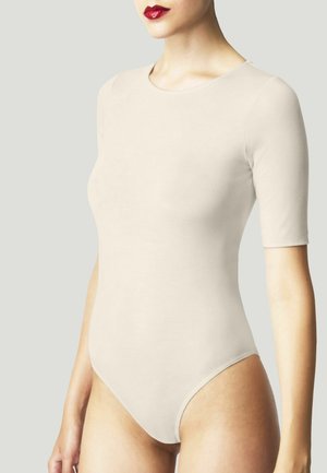 Donna che indossa un body beige aderente con maniche a lunghezza gomito e rossetto rosso acceso su uno sfondo chiaro e uniforme.