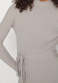 Robe gris clair côtelée avec des manches longues, un col montant et une ceinture nouée à la taille. Le tissu semble doux avec une texture lisse.