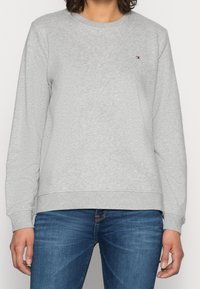 Felpa grigia in cotone con colletto rotondo, maniche lunghe e polsini a coste. Presenta un piccolo logo sul petto. Abbinata a jeans blu.